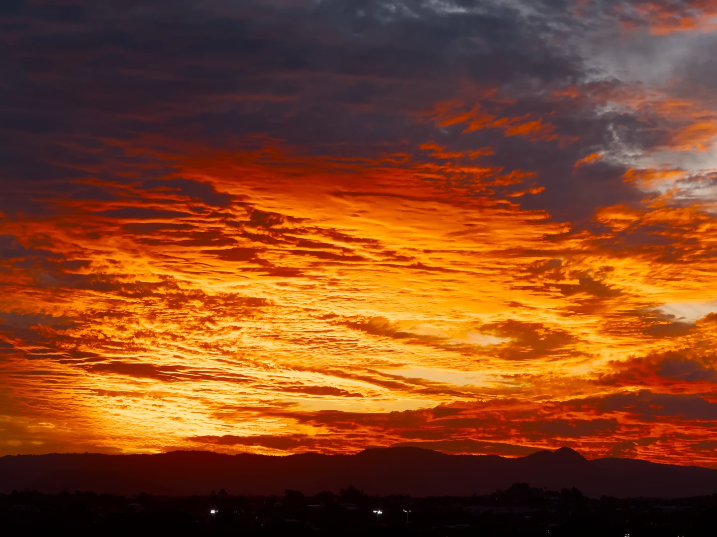 A striking and vivid sunset viewed from Brisbane, looking west over the Taylor Range. The sky fills most of the frame and is covered in layered, wispy clouds lit orange, red, and gold by the setting sun. The upper portion of the sky is darker, with blue-grey cloud cover. The Taylor Range runs across the bottom of the image as a dark silhouette, with two low, rounded peaks visible. A strip of suburban rooftops and a few scattered lights are visible in the foreground shadow below the ridgeline.